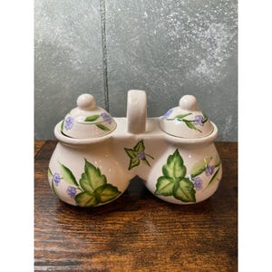 May include: Cream-colored ceramic condiment server with two lidded jars and a handle. The jars are decorated with green leaves and purple flowers. The server is on a wooden surface.