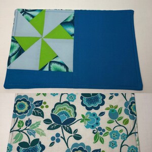 May include: Two blue and green fabric placemats. The top placemat has a blue background with a green and white pinwheel design. The bottom placemat has a white background with a blue and green floral pattern.