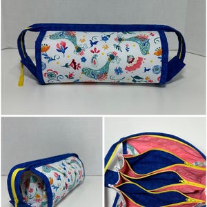 May include: A cylindrical pencil case with a white background, adorned with a colorful floral and bird pattern. The case has a blue trim, handles, and zipper, with a yellow zipper pull. The interior features multiple compartments.