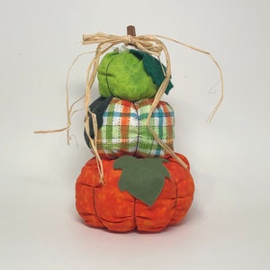 May include: A stack of three fabric pumpkins, the top one is green, the middle one is plaid in green, white, and orange, and the bottom one is orange. The pumpkins are decorated with green fabric leaves.