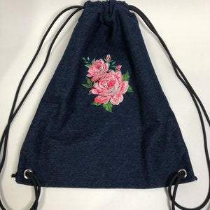 Op de afbeelding: Een donkerblauwe denim trekkoordtas met een roze en groene geborduurde bloemenprint.