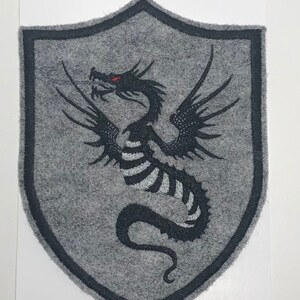 Puede incluir: Un parche de fieltro gris en forma de escudo con un dragón negro bordado con ojos rojos.