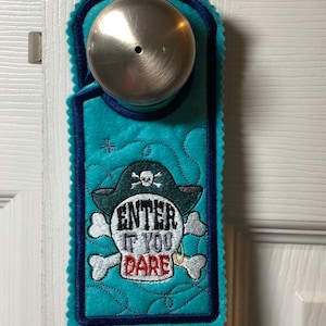 Könnte beinhalten: Ein türhänger in türkisblau mit einem weißen Totenkopf- und Knochenkreuz-Design. Der Totenkopf hat eine schwarze Augenklappe und einen goldenen Ohrring. Der Text "Enter if you dare" ist in weiß und rot auf den Totenkopf gestickt.