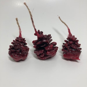 May include: Three red pine cone shaped candles with wicks. The candles are arranged in a row on a white surface.