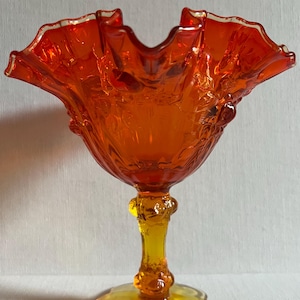 Puede incluir: Un cuenco de pedestal de vidrio naranja con un borde con volantes. El cuenco tiene un patrón floral detallado y un tallo y una base amarillos. El vidrio tiene un acabado brillante y es probablemente vintage.