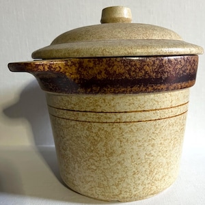 Pode incluir: Uma caçarola de grés vintage com tampa. O prato é bege com salpicos castanhos e uma faixa castanha escura ao redor do meio. Tem uma pega de um lado e duas riscas castanhas. A tampa tem um pequeno botão.