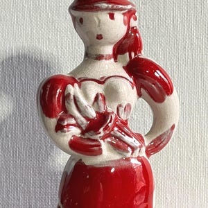 May include: A ceramic figurine of a person wearing a hat and a dress, featuring a glossy red and off-white colour scheme. The figure has a visible crackle glaze finish and is holding a bouquet of flowers in front of the chest area.