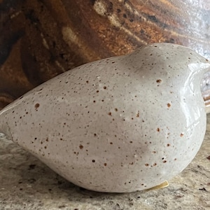 May include: A small, rounded ceramic bird figurine with a glossy, off-white glaze featuring numerous tiny brown speckles across its surface. The bird has a smooth, minimalist shape with a subtle beak and a curved body, showcasing a speckled texture.
