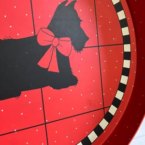 May include: A red metal serving tray with a black silhouette of a dog wearing a red bow. The tray has a red background with white polka dots and a black and white striped border. The tray is round.