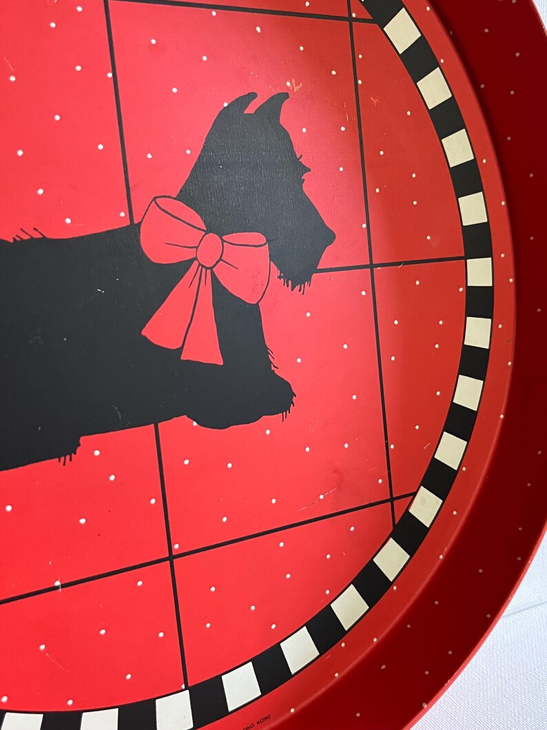 May include: A red metal serving tray with a black silhouette of a dog wearing a red bow. The tray has a red background with white polka dots and a black and white striped border. The tray is round.