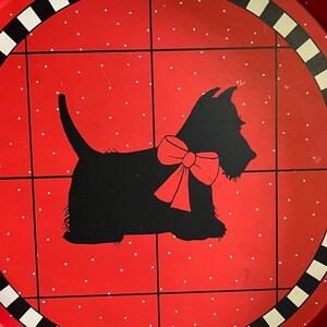 May include: A round red metal tray with a black and white border. The tray features a black silhouette of a Scottish Terrier dog wearing a red bow. The background is red with white polka dots and black grid lines.