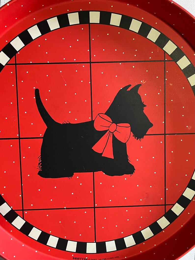 May include: A round red metal tray with a black and white border. The tray features a black silhouette of a Scottish Terrier dog wearing a red bow. The background is red with white polka dots and black grid lines.