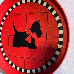 May include: A round red metal tray with a black and white checkered border. The tray features a black silhouette of a dog wearing a red bow, set against a red background with white polka dots. The tray has a vintage aesthetic.