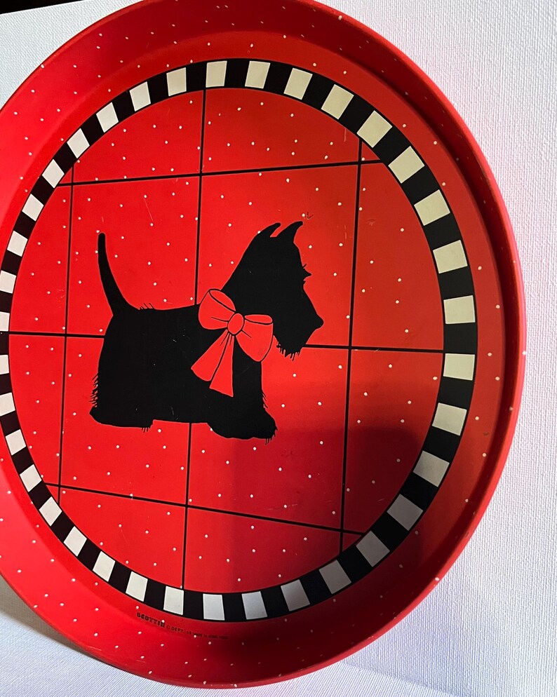 May include: A round red metal tray with a black and white checkered border. The tray features a black silhouette of a dog wearing a red bow, set against a red background with white polka dots. The tray has a vintage aesthetic.