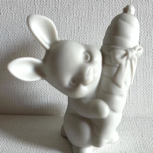 Puede incluir: Figura de conejo de cerámica blanca sosteniendo una zanahoria. El conejo tiene orejas largas y está posado con una ligera inclinación, dándole una apariencia juguetona. La zanahoria está detallada con un lazo.