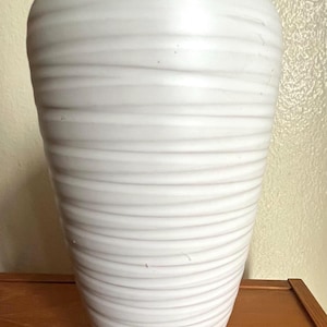 May include: A white ceramic vase with a textured, horizontal ridged design. The vase has a wide top and tapers down to a narrower base. The vase is a solid white color and appears to be handmade.