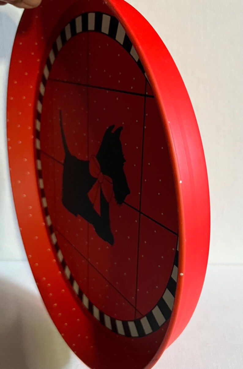 May include: Red, round decorative tray with a black silhouette of a dog wearing a red bow. The tray has a red rim with white polka dots and a black and white striped border.
