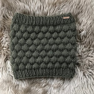 Può includere: Un scaldacollo di lana verde scuro con un motivo testurizzato. Lo scaldacollo ha una piccola etichetta marrone con la parola "naan" su di essa.