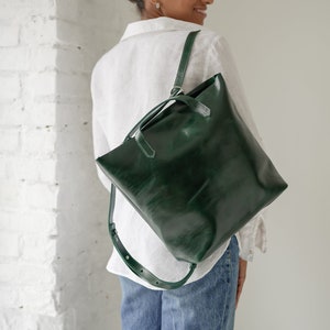 May include: A woman wearing a white shirt and blue jeans is carrying a dark green leather tote bag with a strap that can be worn as a backpack.