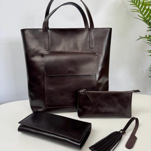 May include: A collection of dark brown leather accessories, including a tote bag with a front pocket, a wallet, a zippered pouch, and a tassel keychain. The items are displayed on a white surface.