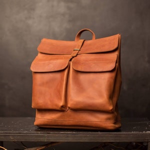 May include: A brown leather backpack with two large front pockets and a flap closure. The backpack has a top handle and adjustable straps.