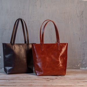 Puede incluir: Dos bolsos de mano de cuero, uno marr&oacute;n y otro marr&oacute;n rojizo, con asas largas, sobre una superficie de madera.