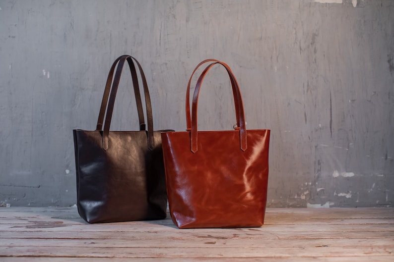 Puede incluir: Dos bolsos de mano de cuero, uno marr&oacute;n y otro marr&oacute;n rojizo, con asas largas, sobre una superficie de madera.