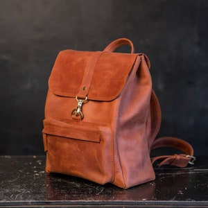 May include: A cognac-colored leather backpack featuring a fold-over flap with a brass-toned metal clasp closure. The bag includes a front zippered pocket, adjustable shoulder straps, and a top handle, showcasing a smooth, matte finish throughout.