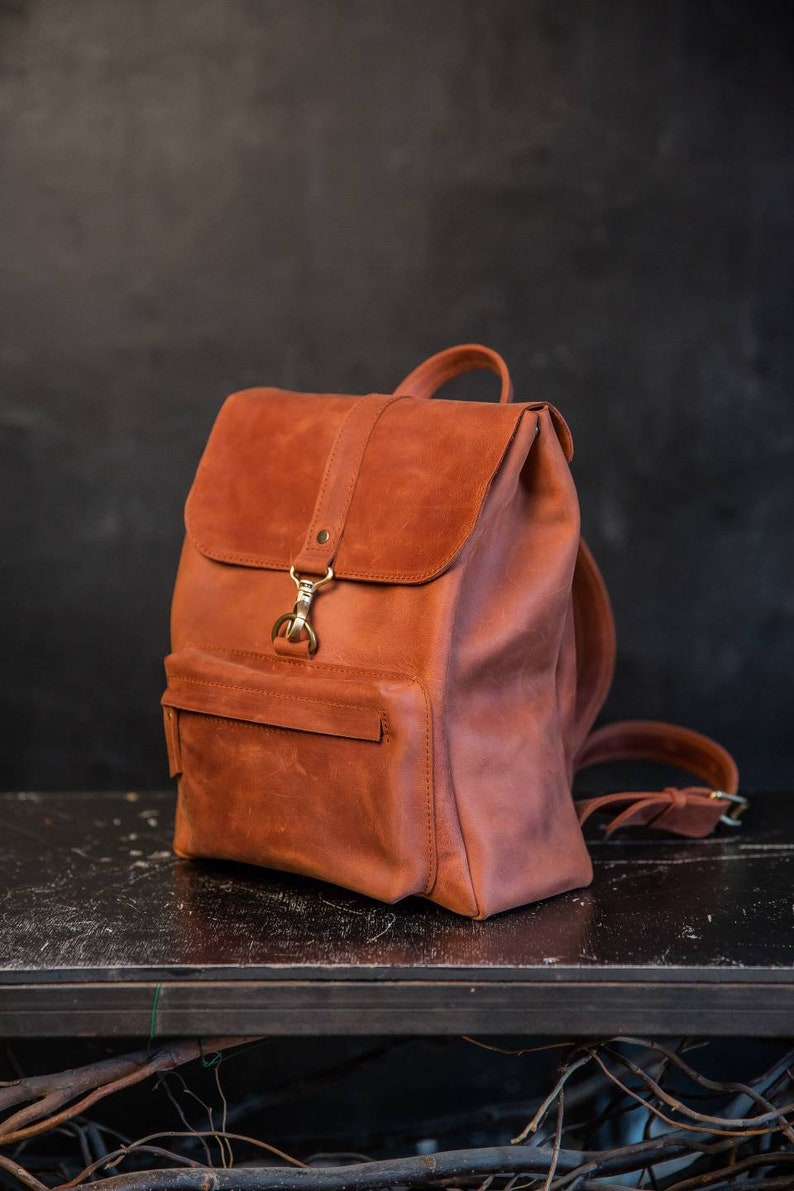 May include: A cognac-colored leather backpack featuring a fold-over flap with a brass-toned metal clasp closure. The bag includes a front zippered pocket, adjustable shoulder straps, and a top handle, showcasing a smooth, matte finish throughout.