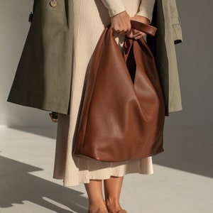 May include: A woman is wearing a beige sweater and skirt with a brown leather tote bag. She is also wearing a green trench coat and brown suede loafers.