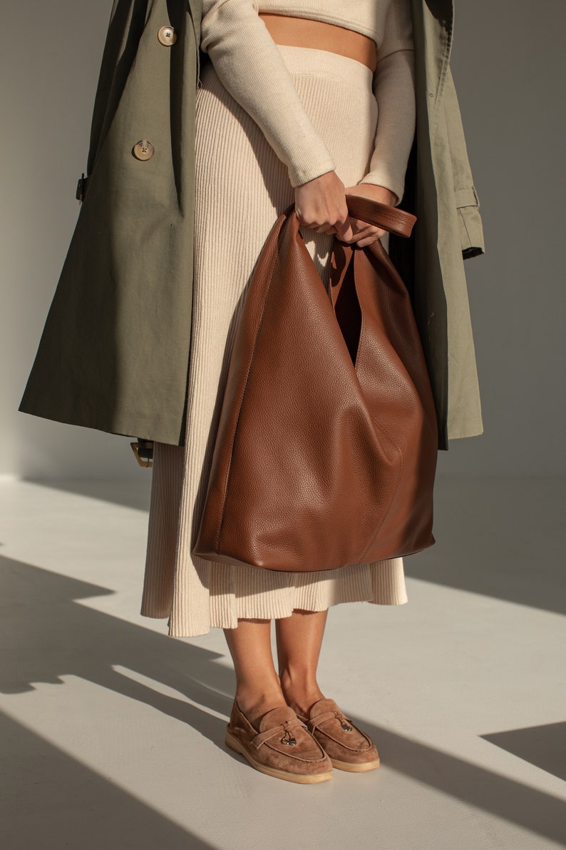 May include: A woman is wearing a beige sweater and skirt with a brown leather tote bag. She is also wearing a green trench coat and brown suede loafers.