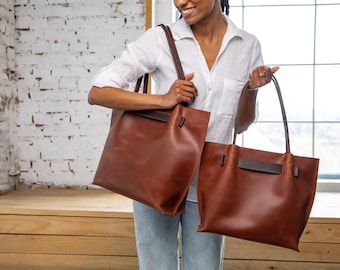 May include: Two brown leather tote bags with dark brown leather handles. The bags are held by a person wearing a white shirt and blue jeans.