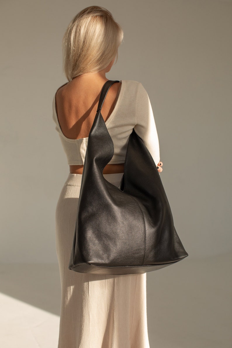 May include: A black leather tote bag with a slouchy design. The bag is worn over the shoulder of a person wearing a white top and skirt.