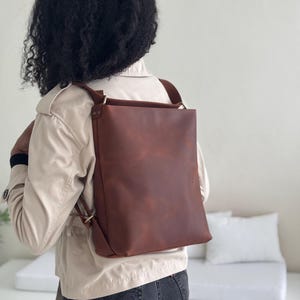 May include: A brown leather backpack with adjustable straps and brass hardware. The bag has a rectangular shape and is worn on the back. The leather has a distressed appearance. The person is wearing a tan jacket and dark jeans.