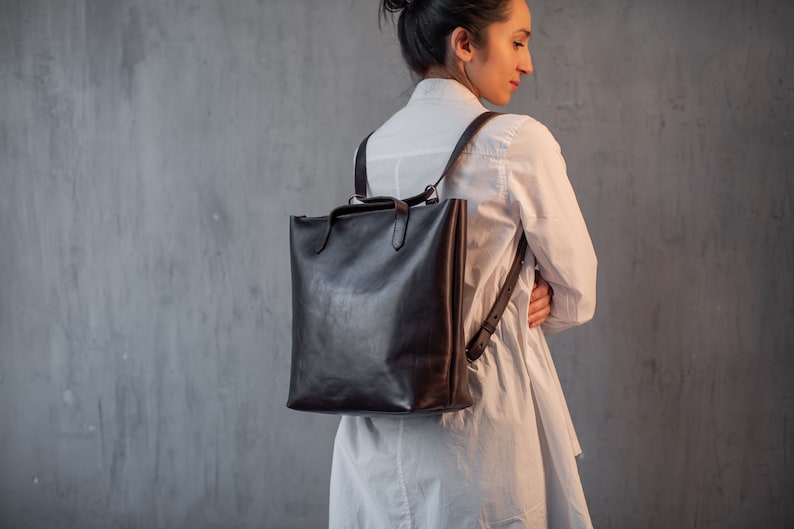 Puede incluir: Un bolso de mano de cuero marr&oacute;n con correas ajustables que se lleva como mochila. El bolso lo lleva una persona que lleva una camisa blanca.