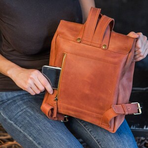 May include: A rust-colored leather backpack featuring a vertical zippered pocket on the back panel, a top handle, and adjustable shoulder straps with metal buckles. A small black electronic device is partially inserted into the zippered pocket.