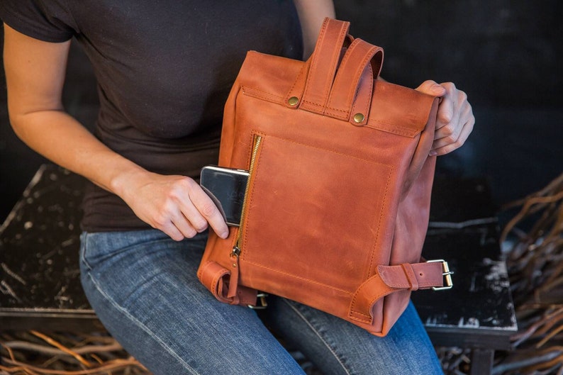 May include: A rust-colored leather backpack featuring a vertical zippered pocket on the back panel, a top handle, and adjustable shoulder straps with metal buckles. A small black electronic device is partially inserted into the zippered pocket.