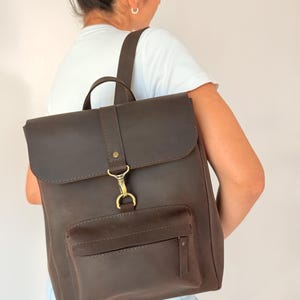 May include: A dark brown leather backpack featuring a rectangular main compartment with a fold-over flap, a front zippered pocket, and a brass-toned metal hook closure. The bag includes adjustable shoulder straps and a top handle for carrying.