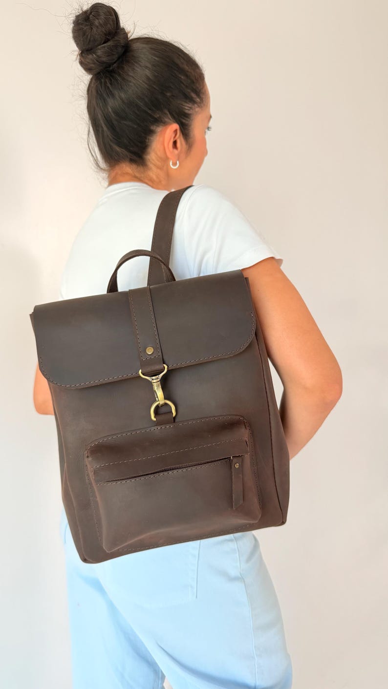 May include: A dark brown leather backpack featuring a rectangular main compartment with a fold-over flap, a front zippered pocket, and a brass-toned metal hook closure. The bag includes adjustable shoulder straps and a top handle for carrying.