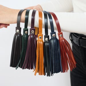 May include: A collection of leather tassels in various colors, including black, green, purple, orange, navy, and brown. Each tassel is attached to a leather strap with a silver-tone rivet. The tassels are held up against a white background.