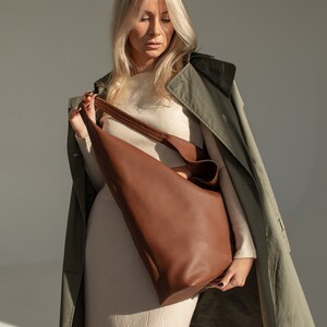 May include: A woman wearing a beige sweater and a green trench coat holds a brown leather tote bag.