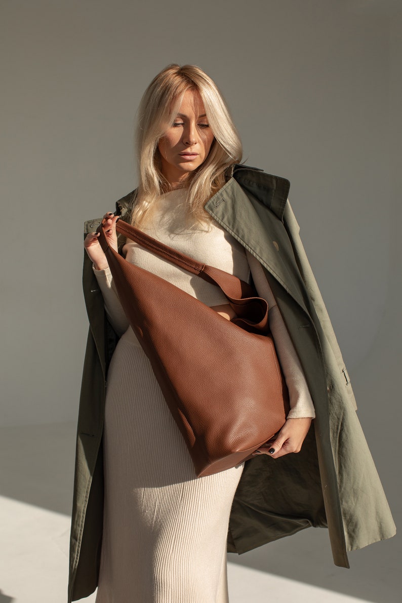 May include: A woman wearing a beige sweater and a green trench coat holds a brown leather tote bag.