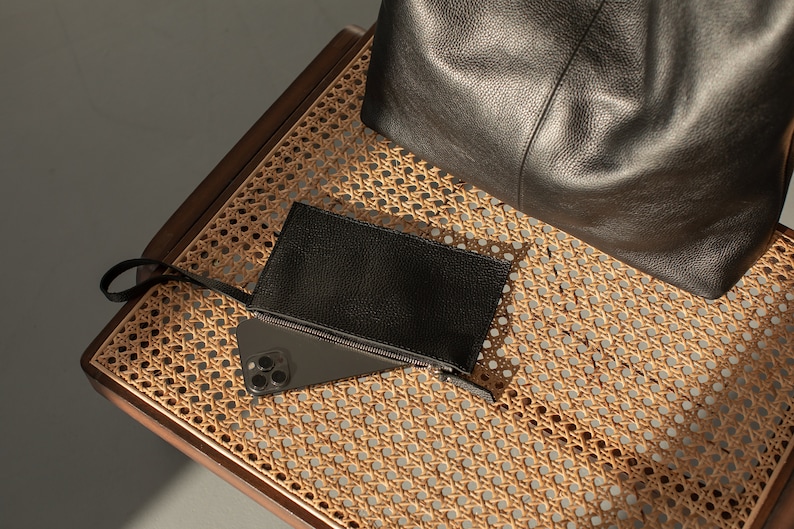 May include: A black leather tote bag and a black leather wallet with a zipper on a woven wicker chair.