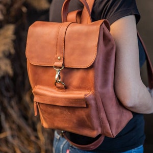 May include: A cognac-colored leather backpack featuring a top handle, adjustable shoulder straps, and a fold-over flap secured with a brass-toned metal lobster clasp. The bag includes a front exterior pocket with a horizontal zipper closure.