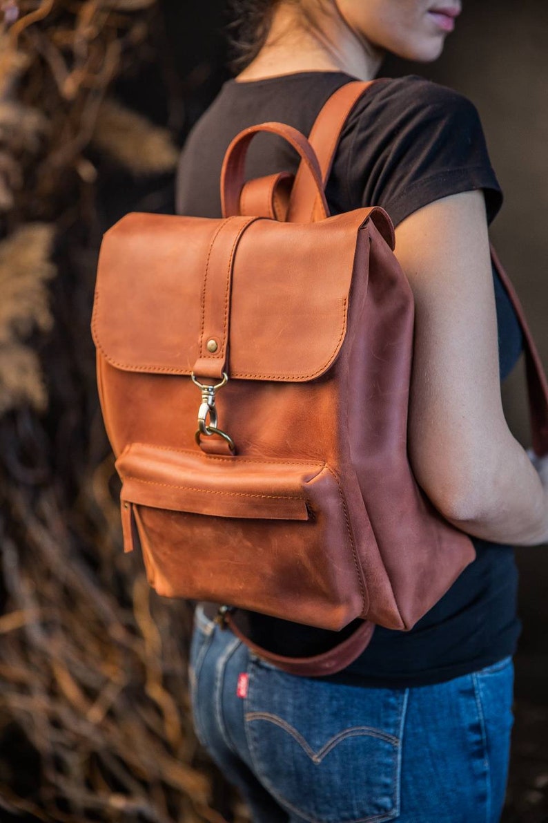 May include: A cognac-colored leather backpack featuring a top handle, adjustable shoulder straps, and a fold-over flap secured with a brass-toned metal lobster clasp. The bag includes a front exterior pocket with a horizontal zipper closure.