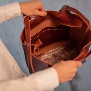 May include: A brown leather tote bag with a zippered pocket and a smaller pocket inside. The bag is open and the interior is visible.