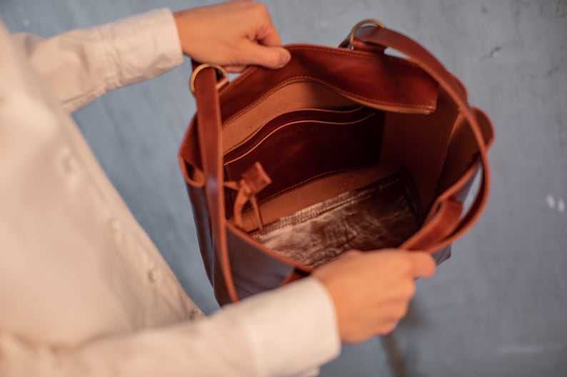 May include: A brown leather tote bag with a zippered pocket and a smaller pocket inside. The bag is open and the interior is visible.