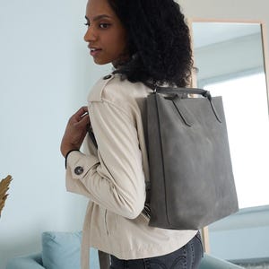 Könnte beinhalten: Eine Frau trägt eine hellbeige Jacke und eine dunkelblaue Jeans und trägt einen grauen Lederrucksack mit verstellbaren Trägern.