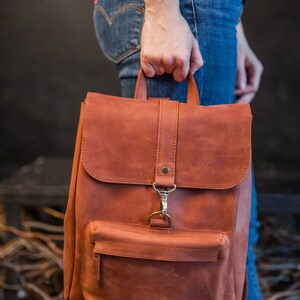 May include: A rust-colored leather backpack featuring a top flap with a vertical strap and a brass-toned metal clasp. The bag includes a front zippered pocket, a top handle, and adjustable shoulder straps with matching brass-toned metal hardware.