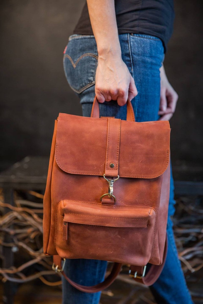 May include: A rust-colored leather backpack featuring a top flap with a vertical strap and a brass-toned metal clasp. The bag includes a front zippered pocket, a top handle, and adjustable shoulder straps with matching brass-toned metal hardware.